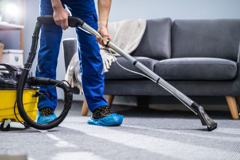 A man in blue pants and shirt vacuums a carpeted room, focusing on cleaning the floor.